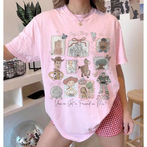 以下が含まれることがあります： トイ・ストーリーのデザインが施されたライトピンクのTシャツ。映画の様々なキャラクターやイメージが特徴で、「Toy Story」と「You've Got a Friend in Me」の文字が入っています。「2026」の年もプリントされています。
