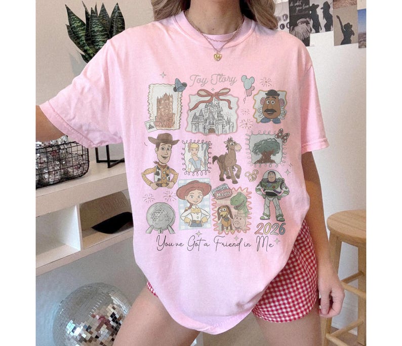 以下が含まれることがあります： トイ・ストーリーのデザインが施されたライトピンクのTシャツ。映画の様々なキャラクターやイメージが特徴で、「Toy Story」と「You've Got a Friend in Me」の文字が入っています。「2026」の年もプリントされています。