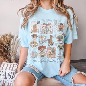 以下が含まれることがあります： 水色のTシャツ。トイ・ストーリーのキャラクターとイメージのコラージュが特徴です。ウッディ、バズ・ライトイヤーなどのイラスト、そして「Toy Story」と「You've Got a Friend in Me」の文字がデザインされています。