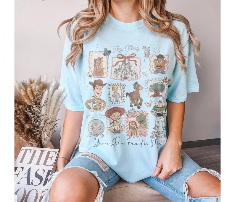 以下が含まれることがあります： 水色のTシャツ。トイ・ストーリーのキャラクターとイメージのコラージュが特徴です。ウッディ、バズ・ライトイヤーなどのイラスト、そして「Toy Story」と「You've Got a Friend in Me」の文字がデザインされています。