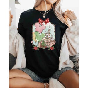 K&ouml;nnte beinhalten: Schwarzes T-Shirt mit Rundhalsausschnitt und einem farbenfrohen Aufdruck eines Schlosses, Luftballons und eines Weihnachtsbaums. Die Grafik enth&auml;lt eine rote Schleife und Mickey Mouse Ohren. Das Design ist mit einem dekorativen Rahmen und roten Weihnachtssternen eingefasst.