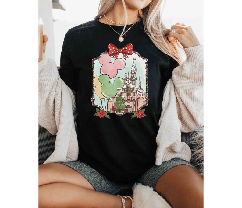 K&ouml;nnte beinhalten: Schwarzes T-Shirt mit Rundhalsausschnitt und einem farbenfrohen Aufdruck eines Schlosses, Luftballons und eines Weihnachtsbaums. Die Grafik enth&auml;lt eine rote Schleife und Mickey Mouse Ohren. Das Design ist mit einem dekorativen Rahmen und roten Weihnachtssternen eingefasst.