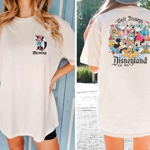 以下が含まれることがあります： ディズニーをテーマにした白いTシャツ2枚。1枚のシャツの前面には、ミニーマウスの漫画の画像と「Mommy」という文字が描かれています。もう1枚のシャツの背面には、ミッキーマウス、ミニーマウス、ドナルドダック、グーフィー、プルート、その他のディズニーキャラクターのカラフルな画像と「Walt Disney's Disneyland Est. 1955」という文字が描かれています。