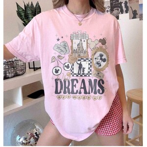 K&ouml;nnte beinhalten: Ein hellrosa T-Shirt mit einem grafischen Design mit einem Schloss, Micky Maus und den Worten "Where Dreams are Made Of". Das Design umfasst Sterne, Kirschen und gerahmte Bilder. Das Shirt wird mit rot-wei&szlig; karierten Shorts getragen.