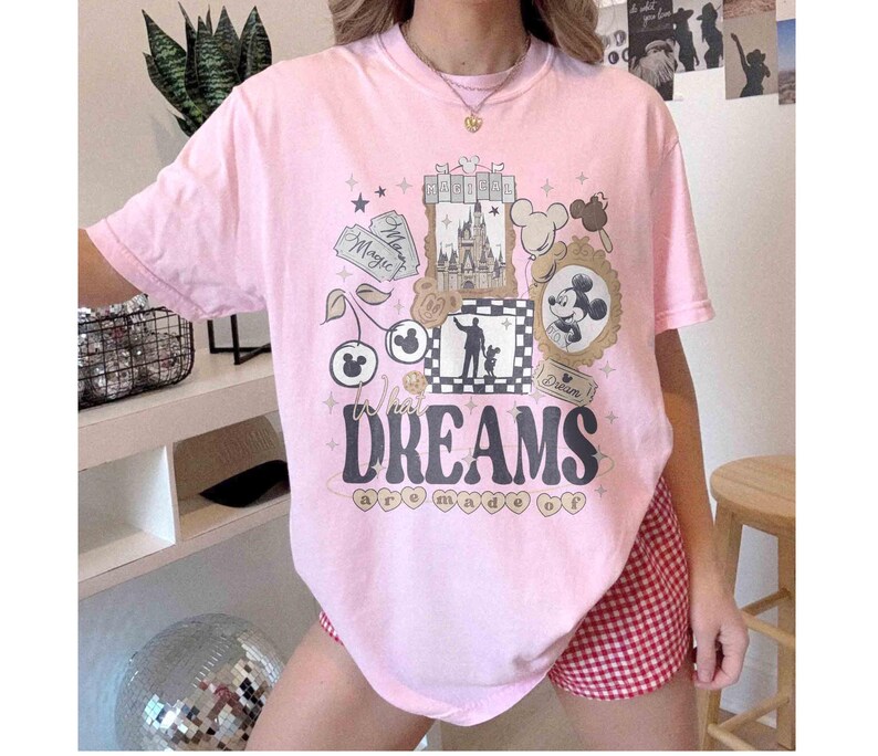 K&ouml;nnte beinhalten: Ein hellrosa T-Shirt mit einem grafischen Design mit einem Schloss, Micky Maus und den Worten "Where Dreams are Made Of". Das Design umfasst Sterne, Kirschen und gerahmte Bilder. Das Shirt wird mit rot-wei&szlig; karierten Shorts getragen.