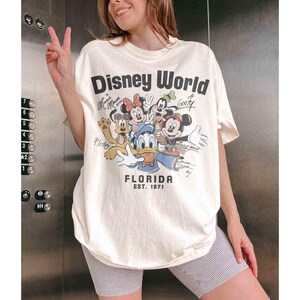 May include: Cream-colored Disney World Florida graphic tee featuring Mickey Mouse, Minnie Mouse, Donald Duck, Goofy, and Pluto. The shirt has the text "Disney World" and "FLORIDA EST. 1971". The shirt is a relaxed fit.