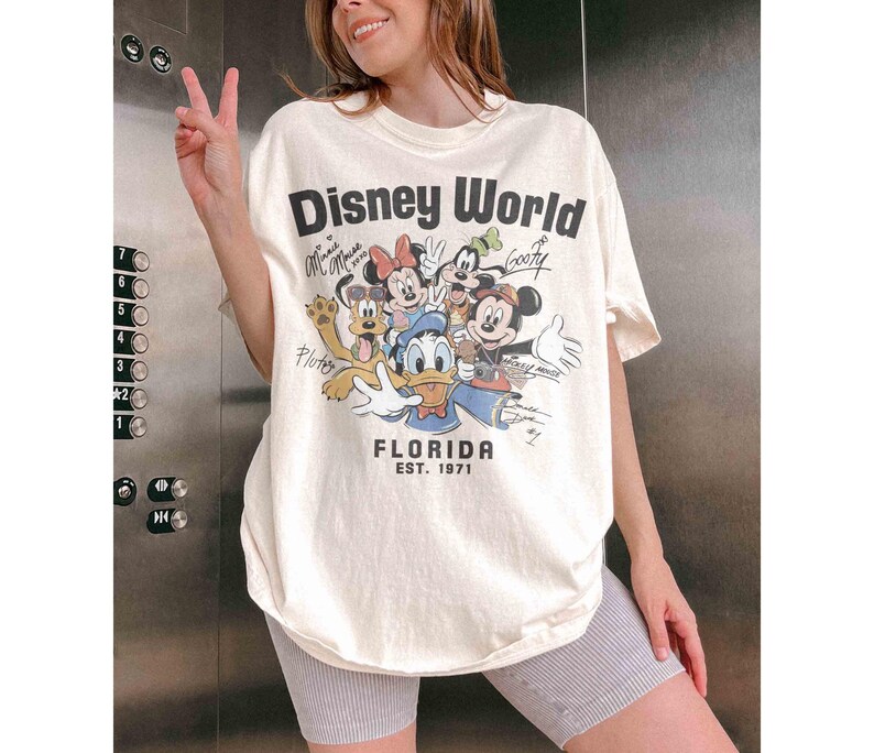 May include: Cream-colored Disney World Florida graphic tee featuring Mickey Mouse, Minnie Mouse, Donald Duck, Goofy, and Pluto. The shirt has the text "Disney World" and "FLORIDA EST. 1971". The shirt is a relaxed fit.