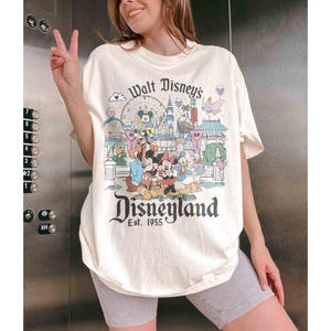May include: Cream-colored Disneyland t-shirt featuring a colorful graphic of Disney characters and park attractions. The shirt has the text "Walt Disney's Disneyland Est. 1955" in a vintage style. The shirt is a relaxed fit.