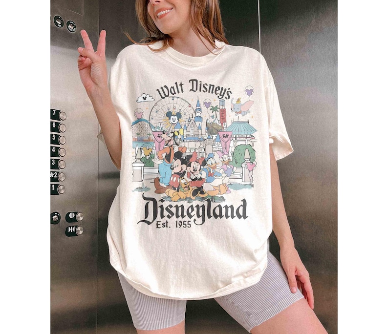 May include: Cream-colored Disneyland t-shirt featuring a colorful graphic of Disney characters and park attractions. The shirt has the text "Walt Disney's Disneyland Est. 1955" in a vintage style. The shirt is a relaxed fit.