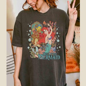 May include: A dark grey t-shirt featuring a vintage-style graphic of Ariel from The Little Mermaid. The graphic depicts Ariel singing with Flounder and Sebastian in the background. The text "The Little Mermaid" is printed below the graphic.
