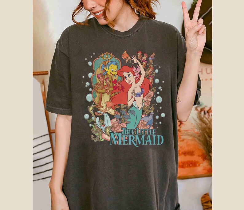 May include: A dark grey t-shirt featuring a vintage-style graphic of Ariel from The Little Mermaid. The graphic depicts Ariel singing with Flounder and Sebastian in the background. The text "The Little Mermaid" is printed below the graphic.