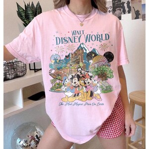 May include: A pale pink t-shirt with a colourful graphic of Disney characters and park attractions. The text "Walt Disney World" is displayed above the illustration, with the phrase "The Most Magical Place On Earth" below.
