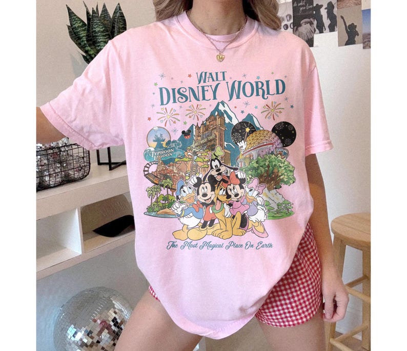 May include: A pale pink t-shirt with a colourful graphic of Disney characters and park attractions. The text "Walt Disney World" is displayed above the illustration, with the phrase "The Most Magical Place On Earth" below.