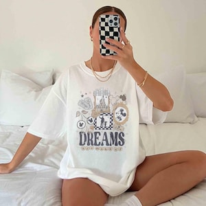 K&ouml;nnte beinhalten: Wei&szlig;es T-Shirt mit einem farbenfrohen Grafikdesign mit dem Wort "DREAMS" und verschiedenen Illustrationen. Das Design enth&auml;lt Bilder eines Schlosses, Mickey Mouse und andere skurrile Elemente. Das Shirt hat eine l&auml;ssige, &uuml;bergro&szlig;e Passform.