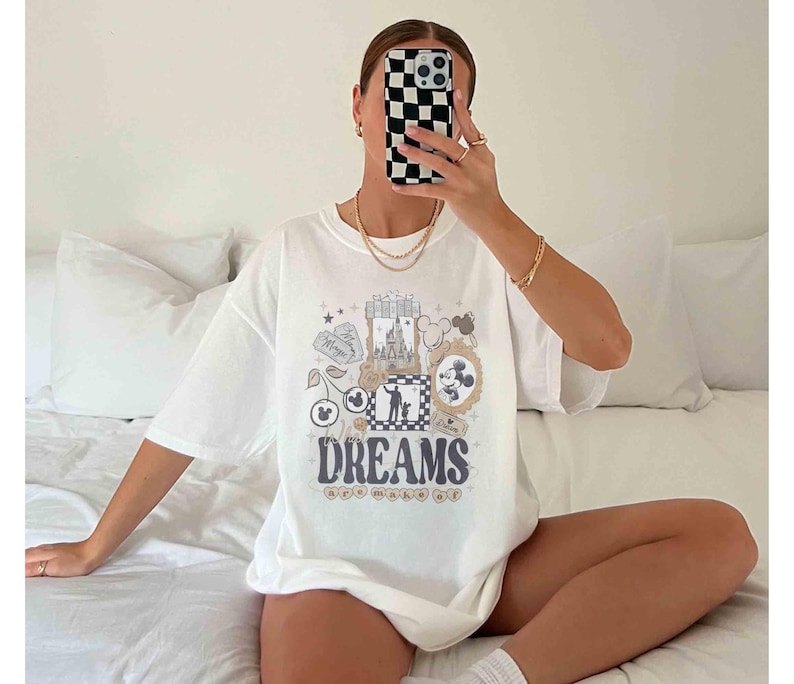K&ouml;nnte beinhalten: Wei&szlig;es T-Shirt mit einem farbenfrohen Grafikdesign mit dem Wort "DREAMS" und verschiedenen Illustrationen. Das Design enth&auml;lt Bilder eines Schlosses, Mickey Mouse und andere skurrile Elemente. Das Shirt hat eine l&auml;ssige, &uuml;bergro&szlig;e Passform.