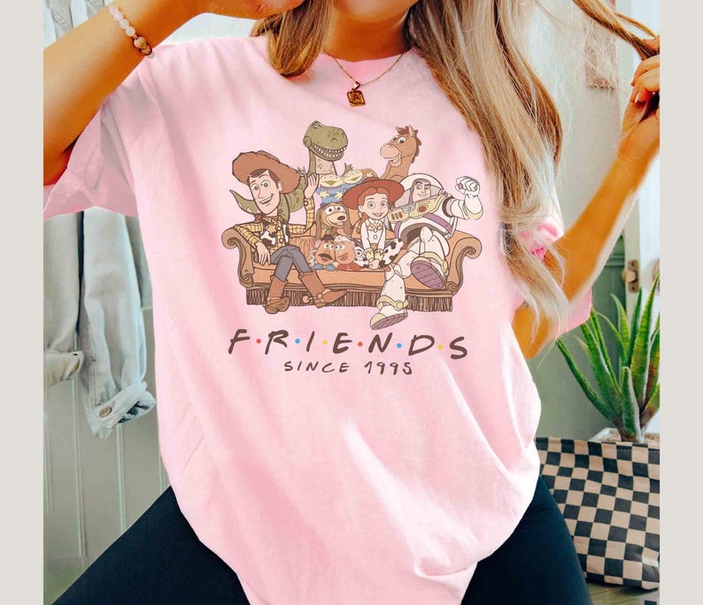 Może przedstawiać: R&oacute;żowa koszulka z grafiką postaci z Toy Story siedzących na kanapie. Pod obrazkiem znajduje się napis "FRIENDS SINCE 1995".