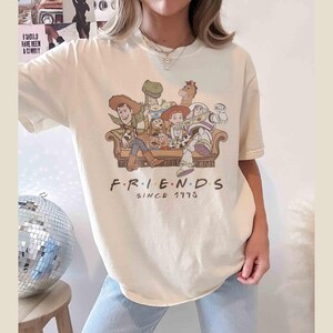 Może przedstawiać: Kremowa koszulka z nadrukiem postaci z Toy Story siedzących na kanapie. Pod obrazkiem znajduje się napis "FRIENDS SINCE 1995".