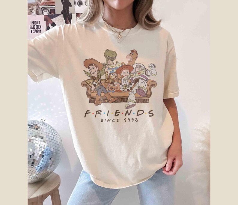 Może przedstawiać: Kremowa koszulka z nadrukiem postaci z Toy Story siedzących na kanapie. Pod obrazkiem znajduje się napis "FRIENDS SINCE 1995".