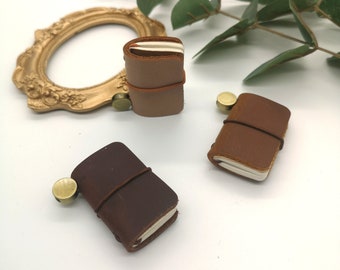 May include: Three miniature leather notebooks in shades of brown, each secured with an elastic band. The notebooks have cream-colored pages and are accompanied by small, round, brass-colored accents. A gold-colored ornate frame and green foliage are in the background.