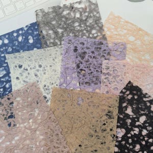 May include: A collection of rectangular sheets of handmade paper featuring a fibrous, web-like texture. The assortment includes various colours such as deep blue, charcoal grey, light peach, lavender, soft pink, tan, and black with visible organic fibres.