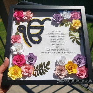 May include: A framed artwork featuring a black and yellow symbol with glitter, surrounded by colourful paper flowers. The artwork includes a text in black, "IK ONKAR SATNAAM KARTA PURAKH NIRBHAU NIRVAIR AKAAL MOORAT AJOONI SAIBHANG GUR PARSAAD JAP AAD SACH JUGAAD SACH HAI BHI SACH NANAK HOSI BHI SACH". The frame is black and the background is white.