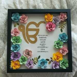 May include: A framed artwork featuring a gold Khanda symbol surrounded by colourful paper flowers. The frame is black and the background is white with a text in Punjabi.