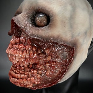May include: A close-up of a white and red latex mask with a grotesque, open-mouthed expression. The mask features a single, large, brown eye and a mouth filled with numerous, sharp teeth.