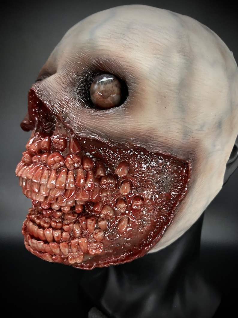 May include: A close-up of a white and red latex mask with a grotesque, open-mouthed expression. The mask features a single, large, brown eye and a mouth filled with numerous, sharp teeth.