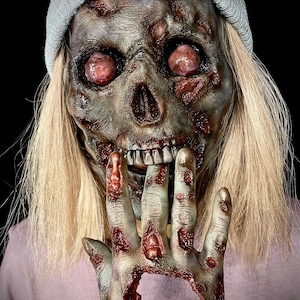 May include: A close-up of a person wearing a grey beanie and a pink shirt. The person's face is covered in a realistic zombie mask with a large open mouth and exposed teeth. The person's hand is also covered in a zombie hand mask with a large hole in the palm.