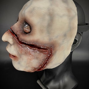 May include: A white latex mask with a realistic, bloody, and gruesome design. The mask features a large gash across the cheek and a visible eye.