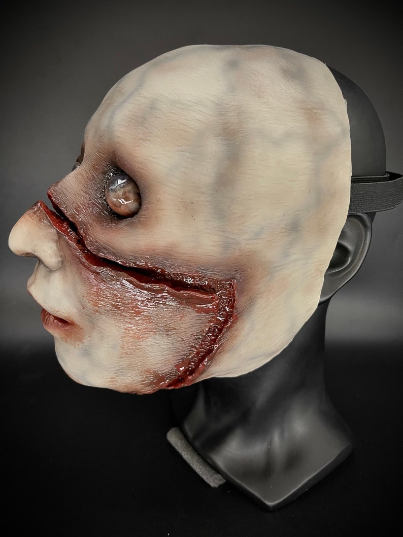 May include: A white latex mask with a realistic, bloody, and gruesome design. The mask features a large gash across the cheek and a visible eye.