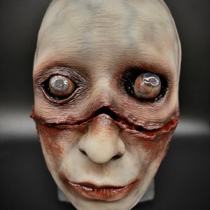May include: A white and grey realistic horror mask with a stitched mouth and bulging eyes. The mask has a red and brown painted effect to give it a bloody and gruesome look.