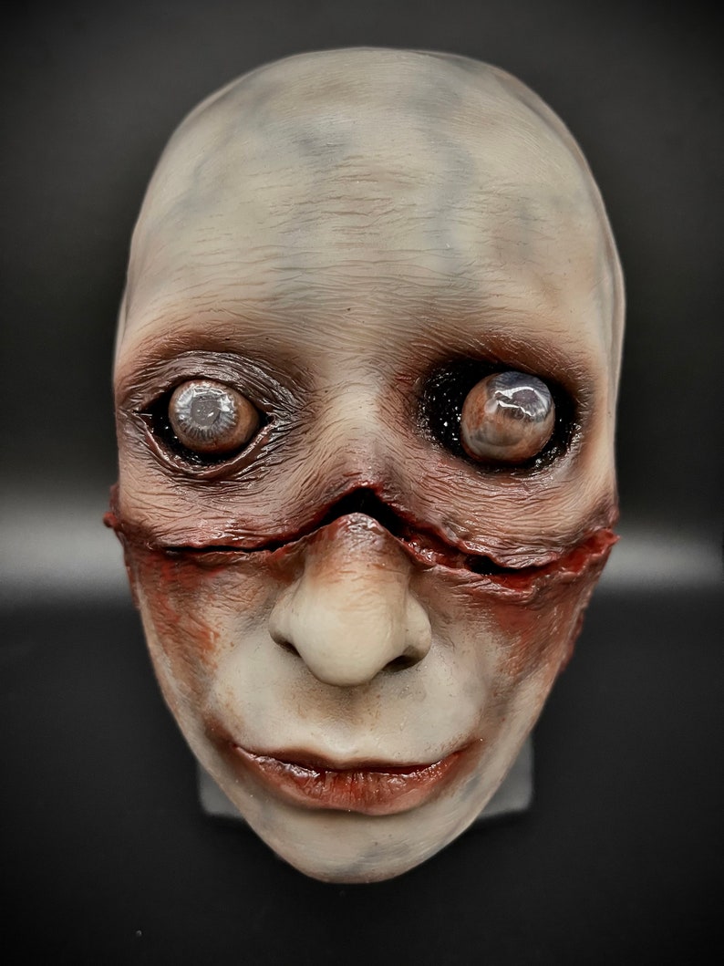 May include: A white and grey realistic horror mask with a stitched mouth and bulging eyes. The mask has a red and brown painted effect to give it a bloody and gruesome look.