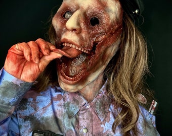 May include: A close-up of a woman wearing a blue and grey uniform with a police badge and the text "BA0093". She is wearing a black police hat and has a grotesque, bloody, and realistic zombie mask on.