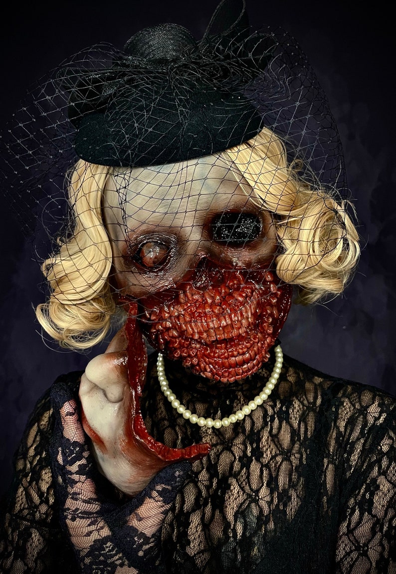 May include: A woman wearing a black lace dress and a black hat with a veil. Her face is covered in gruesome, bloody, and realistic-looking zombie makeup. She is holding a bloody hand in front of her.