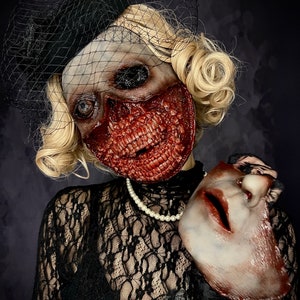 May include: A woman wearing a black lace dress and a black veil holds a white porcelain mask in her right hand. Her face is covered in a gruesome, bloody, and realistic-looking zombie mask.