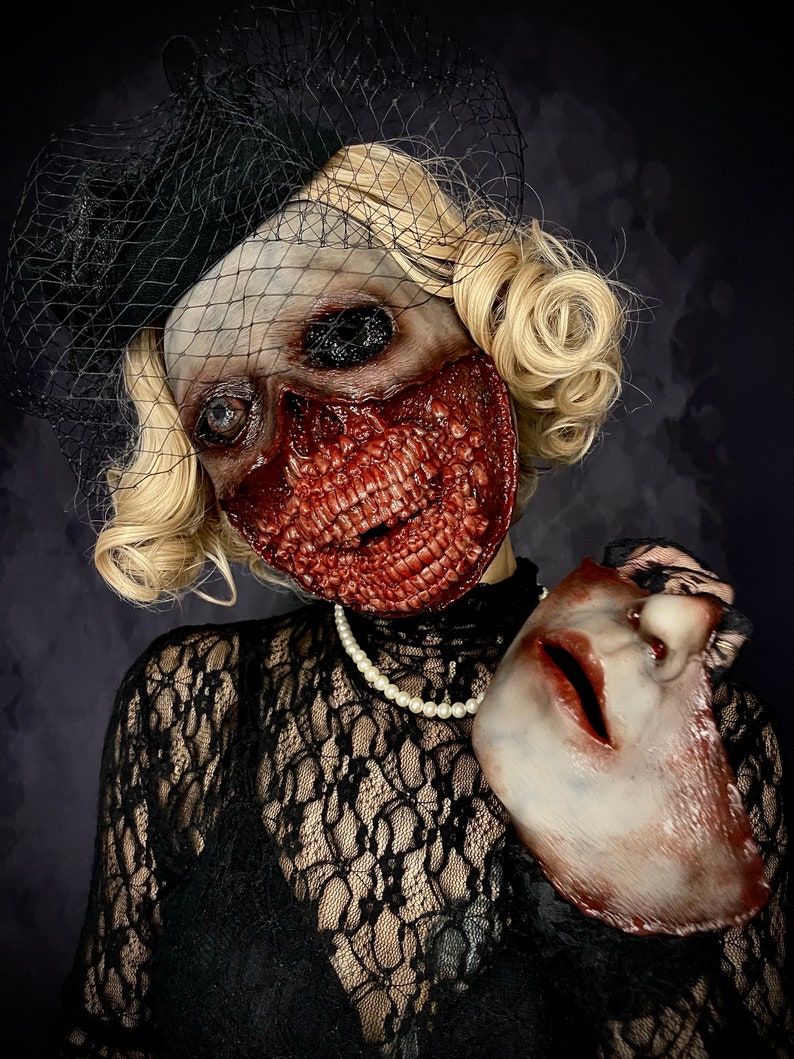 May include: A woman wearing a black lace dress and a black veil holds a white porcelain mask in her right hand. Her face is covered in a gruesome, bloody, and realistic-looking zombie mask.