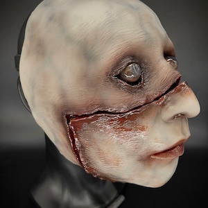 May include: A white latex mask with a realistic, grotesque design. The mask features a large gash across the face with red and brown paint simulating blood and gore. The mask has a realistic eye and a detailed mouth.