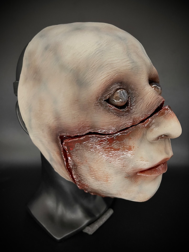 May include: A white latex mask with a realistic, grotesque design. The mask features a large gash across the face with red and brown paint simulating blood and gore. The mask has a realistic eye and a detailed mouth.