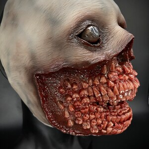 May include: A realistic horror mask featuring a pale, fleshy face and a grotesque, open mouth filled with rows of sharp, white teeth. The mask is made of latex and is designed to be worn over the head.
