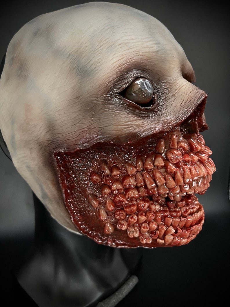 May include: A realistic horror mask featuring a pale, fleshy face and a grotesque, open mouth filled with rows of sharp, white teeth. The mask is made of latex and is designed to be worn over the head.