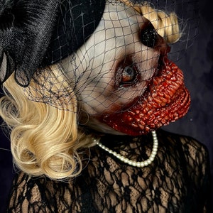 May include: A woman wearing a black lace dress and a black hat with a veil. Her face is covered in a gruesome, bloody makeup effect.