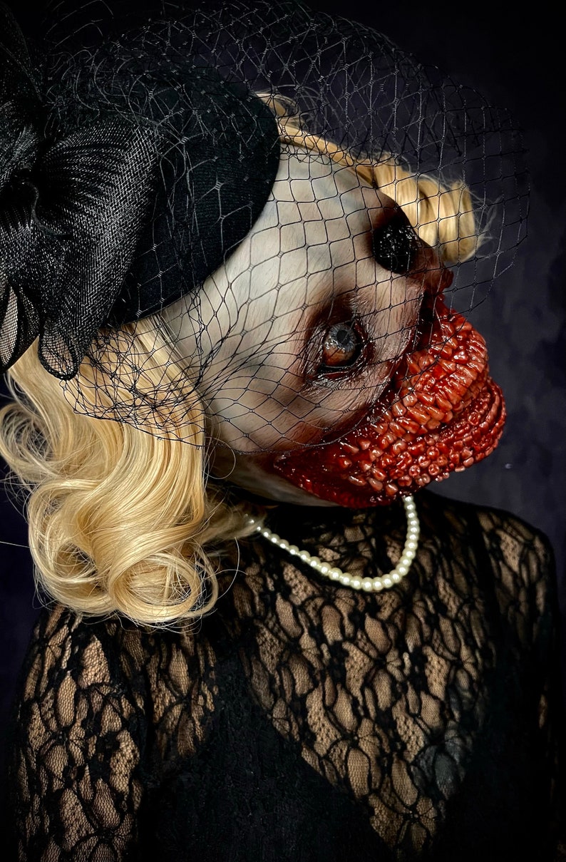 May include: A woman wearing a black lace dress and a black hat with a veil. Her face is covered in a gruesome, bloody makeup effect.
