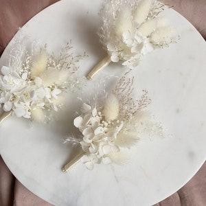May include: Three small bouquets of dried flowers in shades of white and cream. Each bouquet features white hydrangeas, fluffy white pampas grass, and small white flowers. The bouquets are tied with a white ribbon.