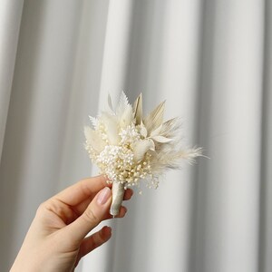 May include: A small bouquet of dried flowers in shades of white and beige, tied with a white ribbon. The bouquet includes dried pampas grass, baby's breath, and other dried flowers.