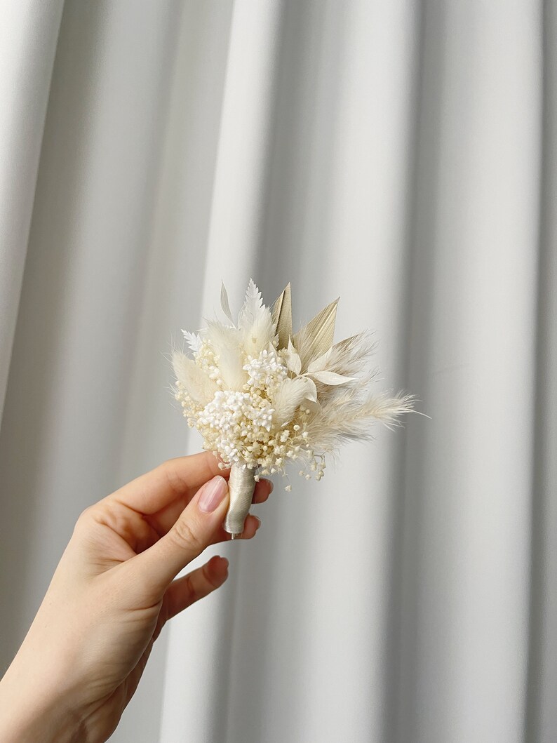 May include: A small bouquet of dried flowers in shades of white and beige, tied with a white ribbon. The bouquet includes dried pampas grass, baby's breath, and other dried flowers.