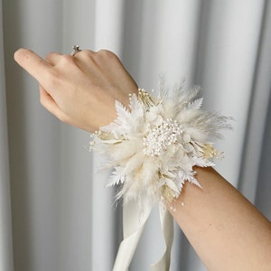 May include: A white floral wrist corsage with a white satin ribbon. The corsage is made of dried flowers and grasses, including pampas grass, baby's breath, and white feathers.