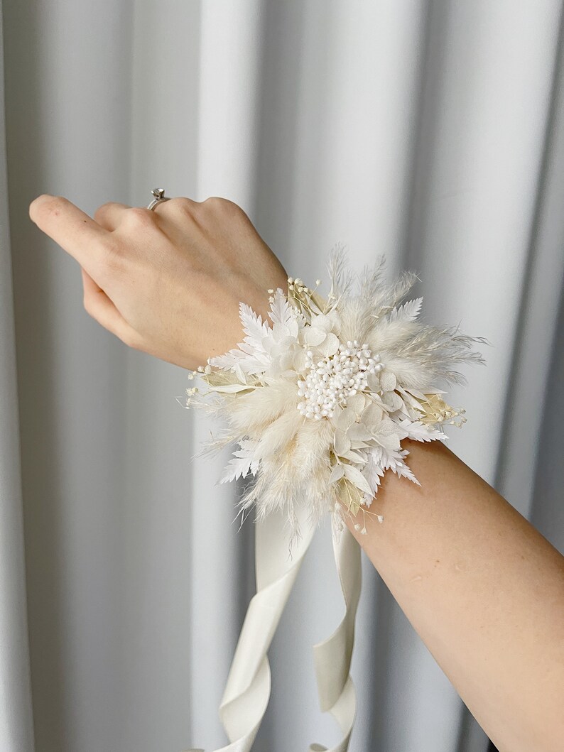 May include: A white floral wrist corsage with a white satin ribbon. The corsage is made of dried flowers and grasses, including pampas grass, baby's breath, and white feathers.