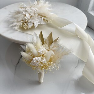 May include: Two white dried flower bouquets with white ribbon. The bouquet on the left has a more traditional flower arrangement, while the bouquet on the right has a more modern, minimalist arrangement.