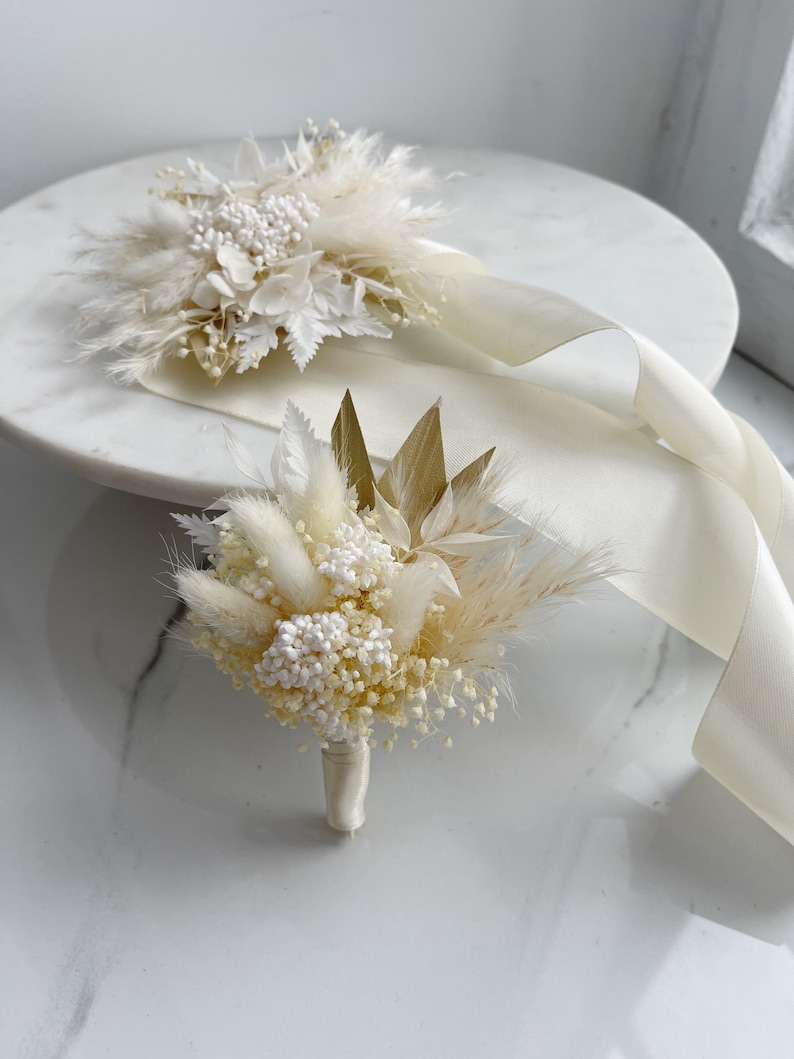 May include: Two white dried flower bouquets with white ribbon. The bouquet on the left has a more traditional flower arrangement, while the bouquet on the right has a more modern, minimalist arrangement.